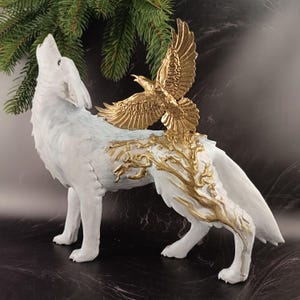 May include: A white wolf figurine with a howling pose, adorned with gold-colored accents, including an eagle and tree-like patterns. The sculpture is detailed and stands on four legs, with a dark background.