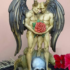 Könnte beinhalten: Eine detaillierte Skulptur von zwei Figuren mit Hörnern und Flügeln, die sich umarmen und eine rote Rose halten. Die Figuren stehen auf einem Sockel mit einem Schädel und Blättern, mit einer Farbpalette aus Gold, Schwarz und Rot.