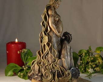 Può includere: Statua dettagliata di una donna incinta con lunghi capelli fluenti, nei toni del beige e del verde. La statua è su una base di legno con la parola "Pachamama" incisa. Una candela rossa e del verde sullo sfondo.