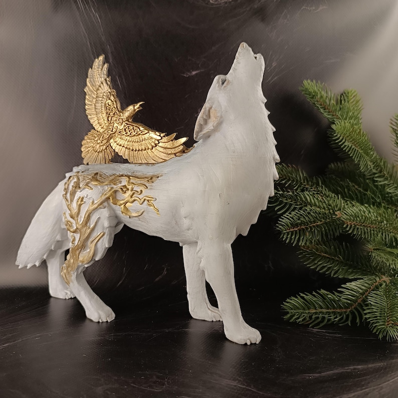 May include: A white wolf figurine with a howling pose, adorned with a gold-colored eagle on its back and gold branch-like accents. The sculpture is set against a dark background with a sprig of evergreen, creating a striking contrast.
