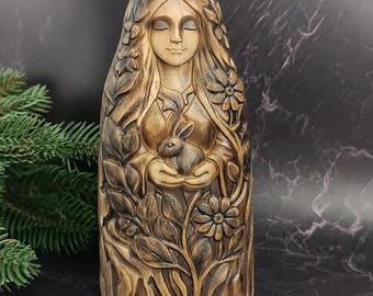 May include: A carved wooden statue of a woman with closed eyes, holding a rabbit. The statue is surrounded by floral and leaf details, with a light brown and dark brown color scheme. The woman's hair is adorned with flowers.