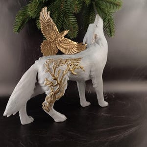 May include: A white wolf figurine with a gold-colored eagle perched on its back. The wolf is in a howling pose, with gold-colored tree-like designs on its side. The eagle has outstretched wings. A sprig of evergreen is in the background.