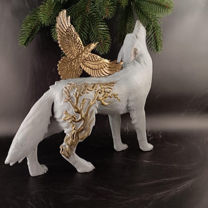 May include: A white wolf figurine with a gold-colored eagle perched on its back. The wolf is in a howling pose, with gold-colored tree-like designs on its side. The eagle has outstretched wings. A sprig of evergreen is in the background.