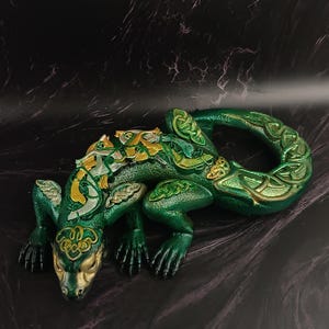 May include: A green and gold lizard figurine with Celtic knot designs. The lizard has a detailed, textured surface and is posed in a relaxed position. The figurine's head and accents are gold, with green and gold patterns along its back and tail.