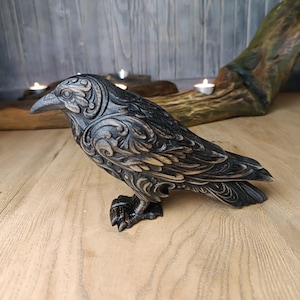 May include: A raven figurine featuring a dark black finish with highlighted bronze-toned edges. The bird is depicted in a standing position with intricate, swirling relief patterns carved across its wings, head, and body, showcasing a detailed texture.