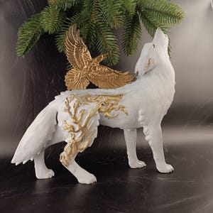 May include: A white wolf figurine with a gold eagle attached to its back. The wolf is in a howling pose, with gold accents on its body. The figurine is a decorative item.