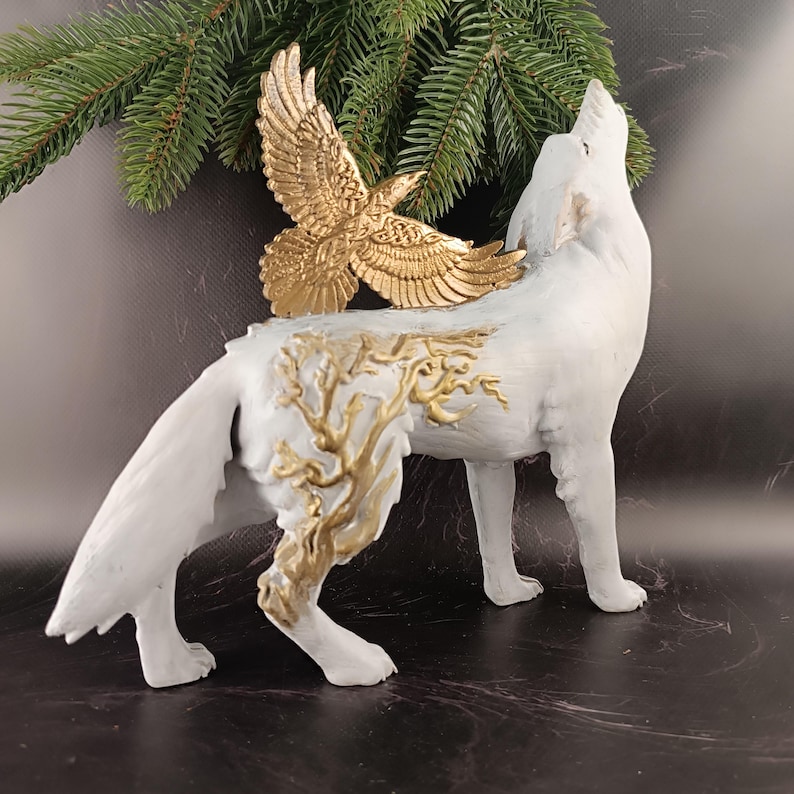 May include: A white wolf figurine with a gold eagle attached to its back. The wolf is howling, and gold-colored tree-like designs adorn its body. A sprig of evergreen is in the background. The figurine is a decorative item.