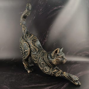 May include: A cat figurine in a stretching pose, featuring a dark charcoal gray base color with intricate, raised gold-toned filigree patterns covering its entire body, including the tail, legs, and head, with a smooth, detailed finish.