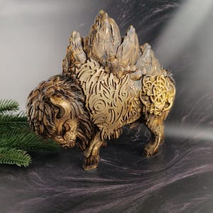 May include: A gold and brown bison figurine featuring intricate carved patterns on its side and a prominent knotwork emblem on its hindquarters. The bison has a textured, rocky hump and a detailed head with curved horns and thick fur.