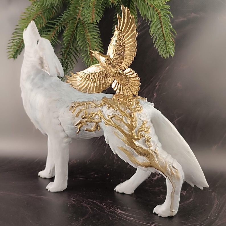 May include: A white wolf figurine with a gold eagle perched on its back. The wolf is howling, and the sculpture features gold tree-like accents on its body. The eagle has outstretched wings, and the sculpture is set against a dark background.