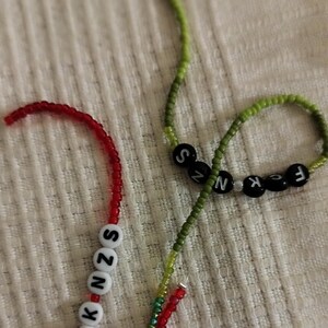 May include: Two beaded necklaces with letter beads spelling out "F*CK NZS". One necklace is red and green, the other is green and black.
