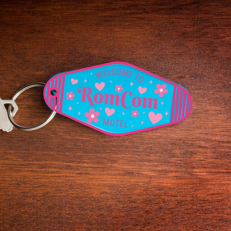May include: A teal and pink motel-style keychain with the words "WELCOME TO RomCom MOTEL" in a retro font. The keychain features pink hearts, flowers, and stripes. A silver key ring is attached to the keychain.