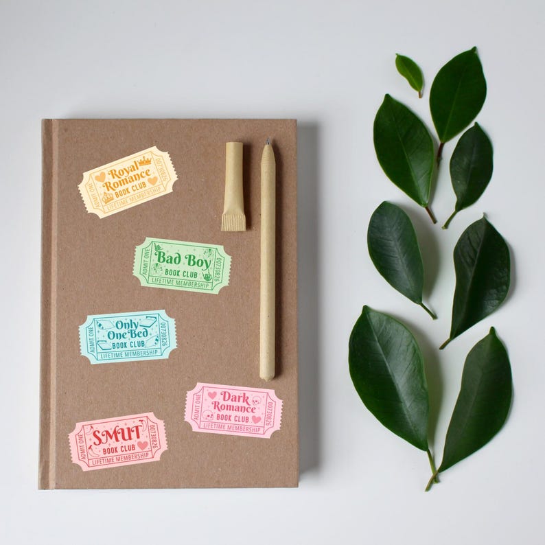 May include: A brown notebook with colorful stickers that read "Royal Romance Book Club," "Bad Boy Book Club," "Only One Bed Book Club," "Smut Book Club," and "Dark Romance Book Club." A pen and a leaf are also present.