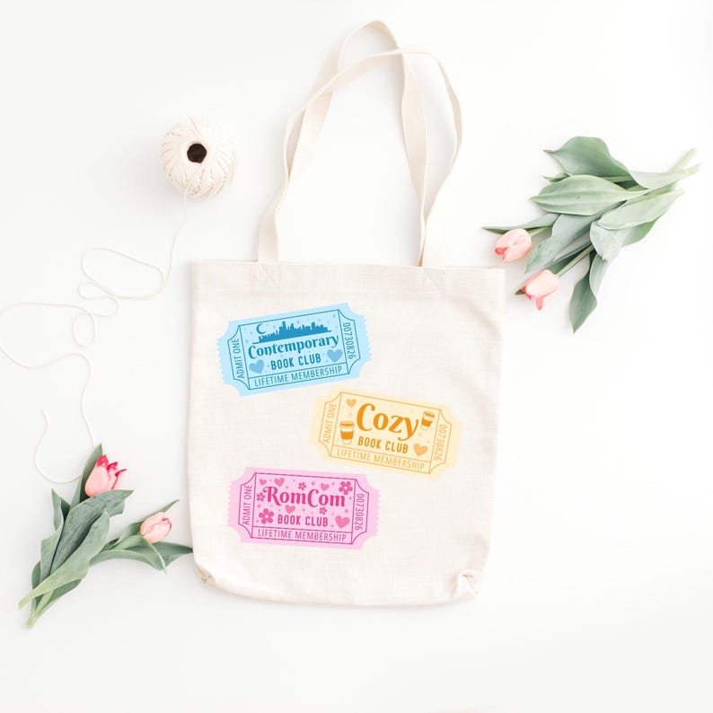 May include: A cream-colored tote bag with three colorful ticket designs. The tickets read "Contemporary Book Club," "Cozy Book Club," and "RomCom Book Club." The bag is decorated with pink and green flowers and a ball of twine.