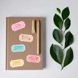 May include: A brown notebook with colorful ticket-shaped stickers that read "Pining/Yearning Book Club," "Magic School Book Club," "Fantasy Romance Book Club," "Royal Romance Book Club," and "Cursed Lovers Book Club." A pen and a pen holder are on the notebook, with green leaves on the side.