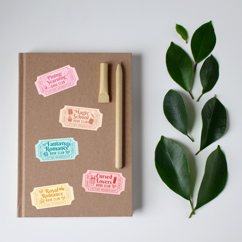 May include: A brown notebook with colorful ticket-shaped stickers that read "Pining/Yearning Book Club," "Magic School Book Club," "Fantasy Romance Book Club," "Royal Romance Book Club," and "Cursed Lovers Book Club." A pen and a pen holder are on the notebook, with green leaves on the side.