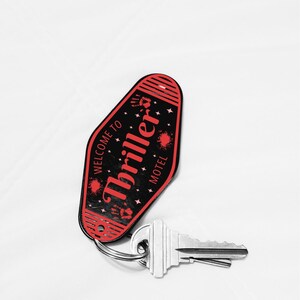 May include: A black and red motel key tag with the words "Thriller Motel" and "Welcome to" printed on it. The key tag has a key ring attached to it, and a silver key is connected to the ring. The key tag has a retro design with star and handprint graphics.