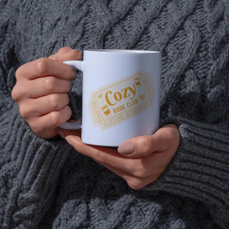 May include: A white ceramic mug featuring a gold ticket design that reads "Cozy Book Club Lifetime Membership." The design includes coffee cup illustrations and heart accents. The mug is held against a gray cable-knit sweater.