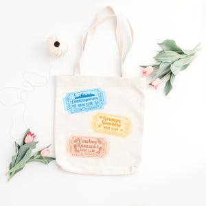 May include: A cream-colored tote bag with three colorful ticket-shaped designs. The tickets read "Contemporary Book Club," "Grumpy Sunshine Book Club," and "Cowboy Romance Book Club." The bag has a simple design, perfect for carrying books.