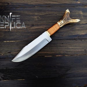 May include: A hunting knife with a brown leather handle and a stag antler pommel. The blade is silver and has a slight curve. The knife is lying on a dark wood surface.