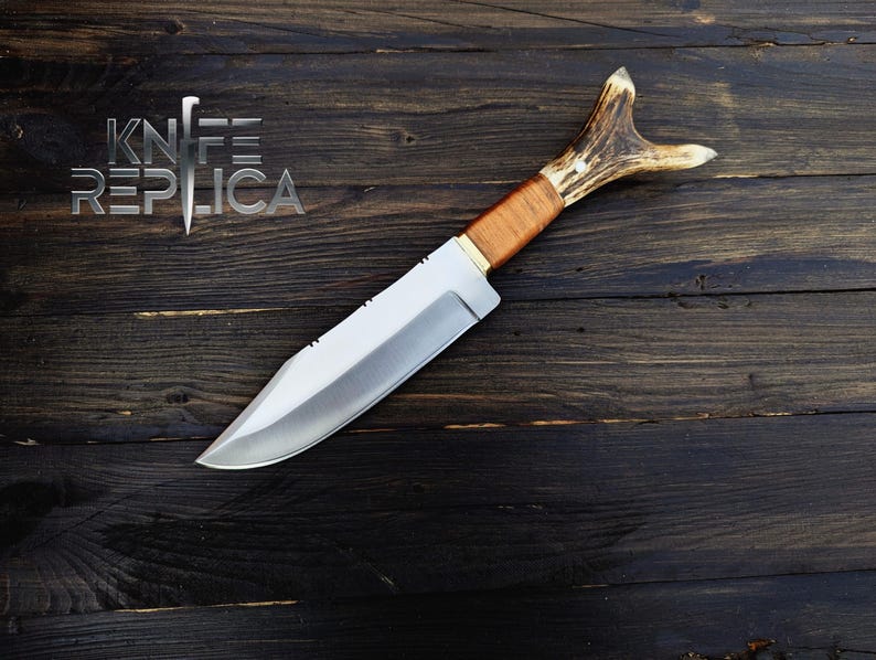 May include: A hunting knife with a brown leather handle and a stag antler pommel. The blade is silver and has a slight curve. The knife is lying on a dark wood surface.