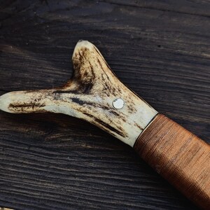 May include: A brown leather wrapped handle with a white antler pommel. The antler has a small metal ring embedded in it.