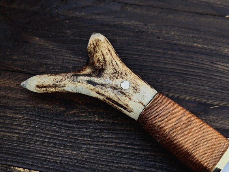 May include: A brown leather wrapped handle with a white antler pommel. The antler has a small metal ring embedded in it.