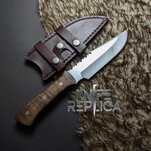 May include: A hunting knife with a wooden handle and a serrated blade, accompanied by a brown leather sheath. The knife has a shiny, silver blade and is set against a textured, brown background. The text "KNIFE REPLICA" is visible.