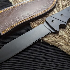 May include: A black knife with a serrated edge and a dark handle. The knife is partially sheathed in a brown leather case with decorative stitching. The blade is angled towards the viewer.