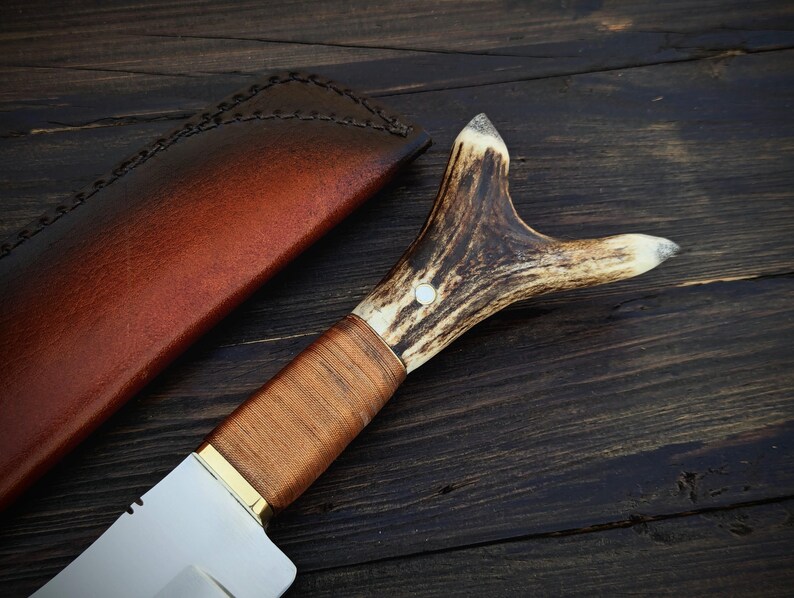 May include: A hunting knife with a brown leather sheath and a stag antler handle. The blade is silver and has a slight curve.