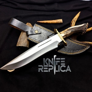 May include: A large hunting knife with a polished silver blade and a handle made of deer antler. The knife has a brass guard and is accompanied by a black leather sheath. The text "KNIFE REPLICA" is visible. The knife is displayed on a black surface.