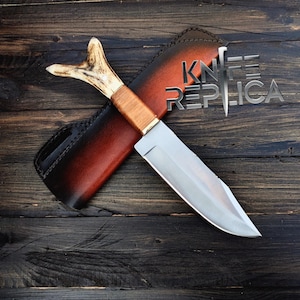 May include: A hunting knife with a brown wooden handle and a silver blade. The knife is in a brown leather sheath with a red accent. The sheath has the text "KNIFE REPLICA" on it.