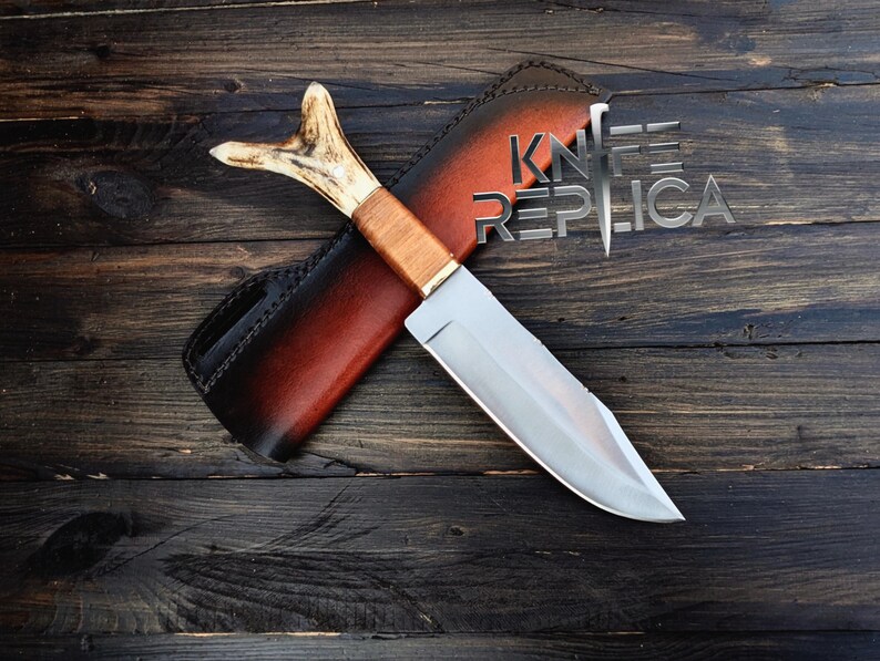 May include: A hunting knife with a brown wooden handle and a silver blade. The knife is in a brown leather sheath with a red accent. The sheath has the text "KNIFE REPLICA" on it.