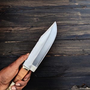May include: A silver hunting knife with a brown antler handle. The knife is held in a hand against a dark wood background.