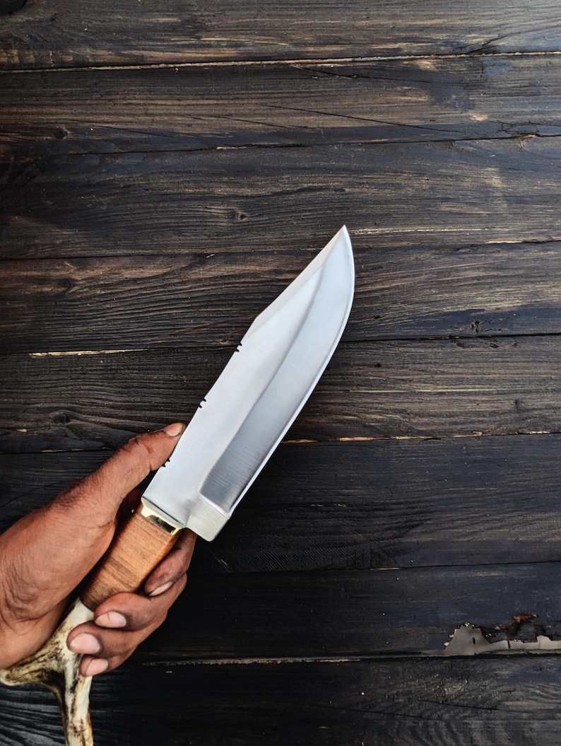 May include: A silver hunting knife with a brown antler handle. The knife is held in a hand against a dark wood background.