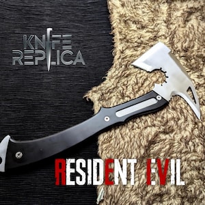 Pode incluir: Um machado de metal e preto com uma lâmina prateada e um cabo preto. O texto "KNIFE REPLICA" está acima do machado, e o texto "RESIDENT EVIL" abaixo.
