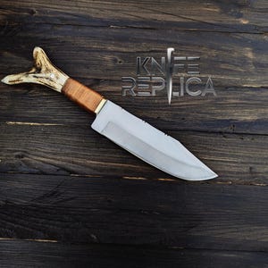 May include: A hunting knife with a brown leather handle and a white antler pommel. The blade is silver and has a slight curve. The knife is lying on a dark wooden surface.