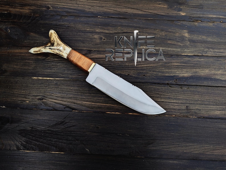 May include: A hunting knife with a brown leather handle and a white antler pommel. The blade is silver and has a slight curve. The knife is lying on a dark wooden surface.