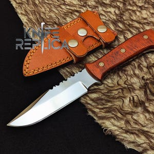May include: A hunting knife with a wooden handle and a serrated blade, accompanied by a brown leather sheath. The knife features a polished steel blade and is secured in the sheath with a snap closure. The handle is a reddish-brown color.