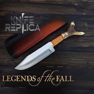 May include: A hunting knife with a brown leather sheath and a stag antler handle. The knife has a silver blade and is on a dark wood background. The text "Legends of the Fall" is written in gold lettering below the knife.