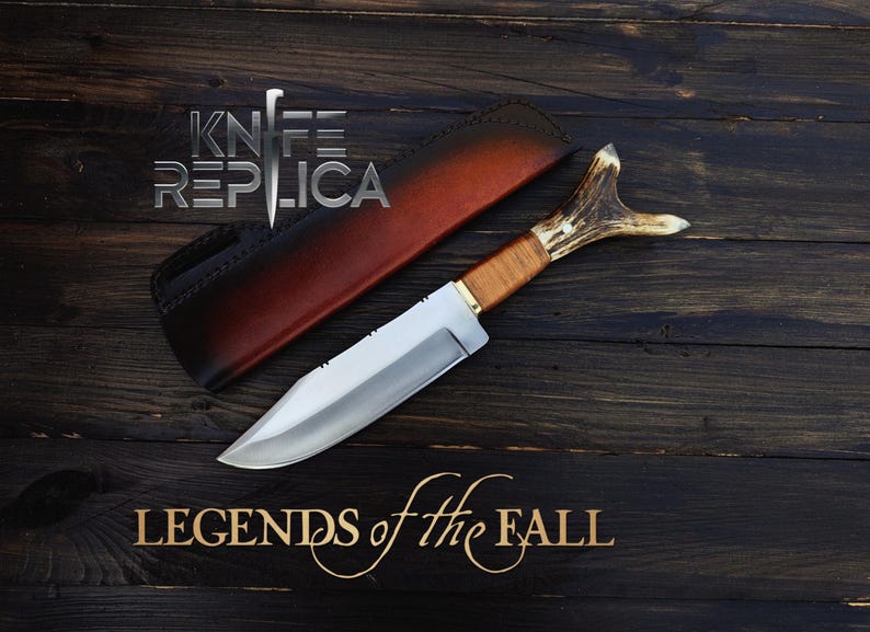 May include: A hunting knife with a brown leather sheath and a stag antler handle. The knife has a silver blade and is on a dark wood background. The text "Legends of the Fall" is written in gold lettering below the knife.