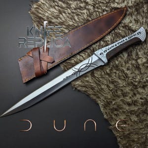 May include: A silver dagger with an ornate handle and a brown leather sheath. The blade features etched designs, and the handle has a dark wood grain. The sheath is secured with a leather strap. The text "KNIFE REPLICA" is visible.