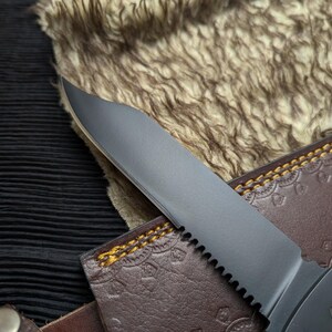 May include: Close-up of a black serrated knife blade with a brown leather sheath. The sheath has a decorative embossed pattern and yellow stitching. The knife blade is partially visible, with a sharp edge and a dark finish.