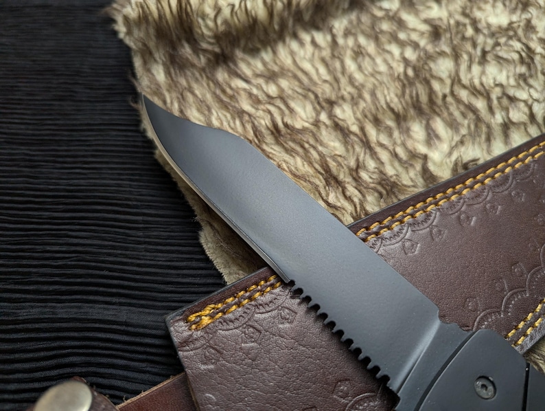 May include: Close-up of a black serrated knife blade with a brown leather sheath. The sheath has a decorative embossed pattern and yellow stitching. The knife blade is partially visible, with a sharp edge and a dark finish.