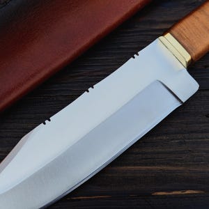 May include: A silver-colored hunting knife with a brown leather handle and a brass accent. The knife is lying on a dark wood surface.