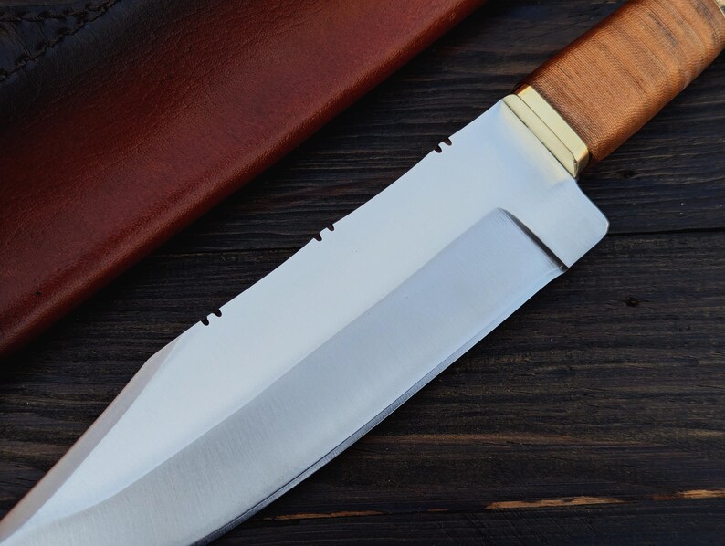 May include: A silver-colored hunting knife with a brown leather handle and a brass accent. The knife is lying on a dark wood surface.