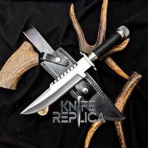 May include: A large, silver-bladed knife with a black handle and a black leather sheath. The knife is displayed on a black surface with deer antlers. The text "KNIFE REPLICA" is visible.