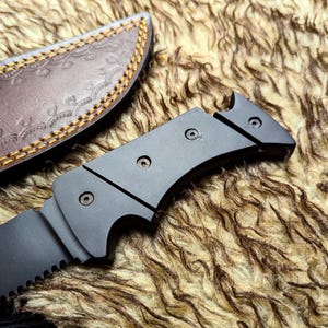 May include: A black survival knife with a serrated blade and a dark handle. The knife is partially visible next to a brown leather sheath with decorative stitching.