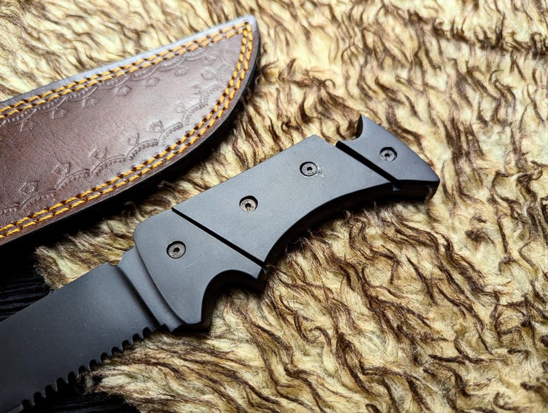 May include: A black survival knife with a serrated blade and a dark handle. The knife is partially visible next to a brown leather sheath with decorative stitching.