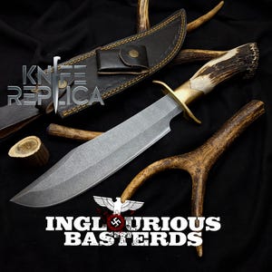 May include: A large,  handmade knife with a brown leather sheath and a stag antler handle. The blade is made of steel and has a textured finish. The knife is displayed on a black background with antler pieces. The text "INGLOURIOUS BASTERDS" is visible in the lower part of the image.